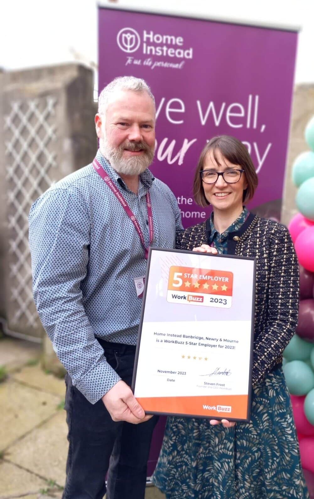 Two people holding a framed 5 Star Employer certificate, standing in front of a Home Instead banner and colorful balloons. - Home Instead