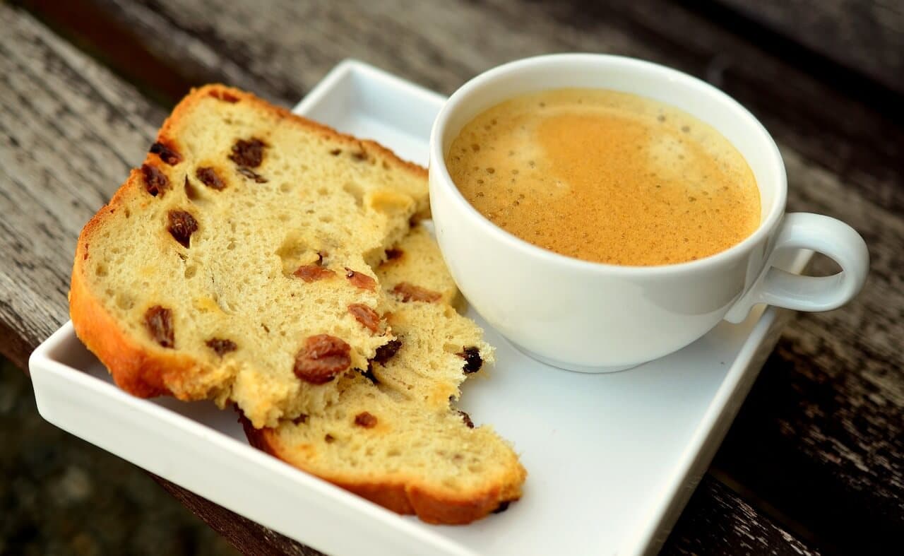 A white cup of coffee and two slices of fruit bread on a white square plate, placed on a wooden surface. - Home Instead