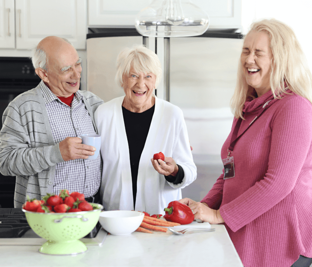 Three people, an elderly couple and their carer, laughing together in a kitchen as the carer prepares fruit and vegetables for a meal