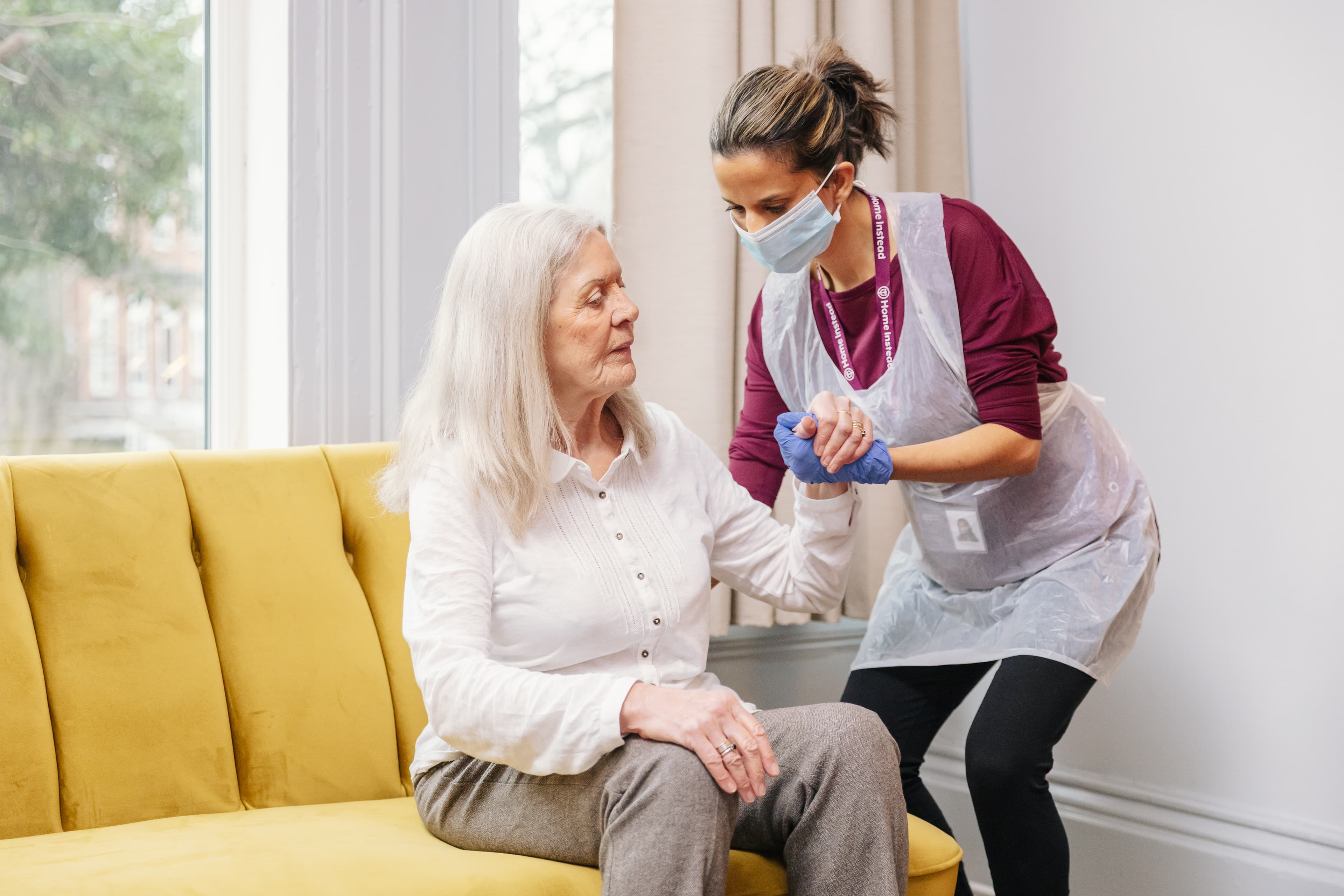 Caregiver helping an elderly woman stand up from a yellow couch, both wearing masks and protective gear. - Home Instead