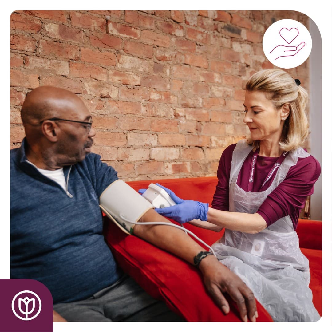 A healthcare worker checks an older man's blood pressure in a cozy room with a brick wall. Health icon in the corner. - Home Instead