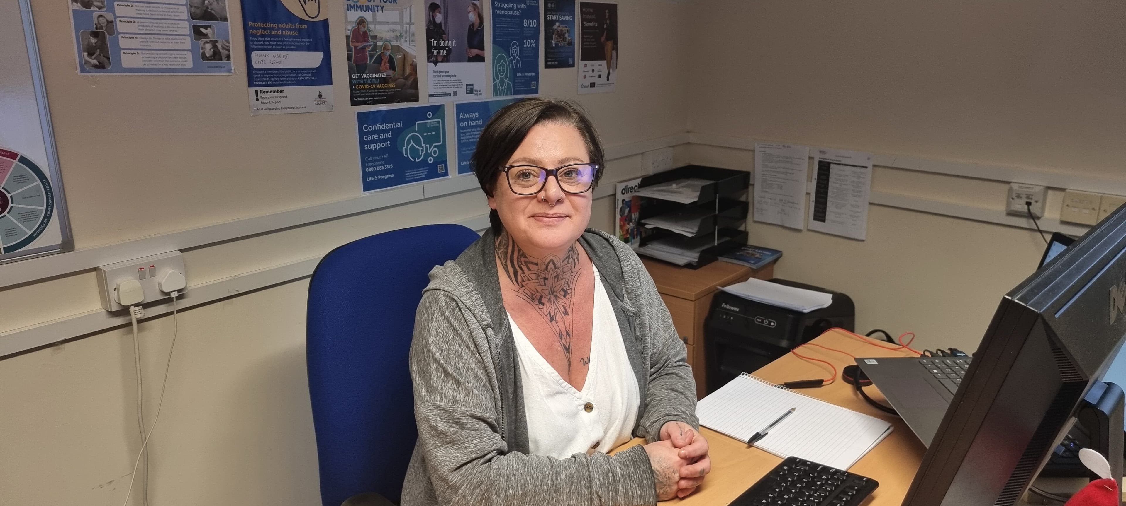 Woman with glasses and neck tattoos sits at a desk in an office with posters on the wall behind her. - Home Instead