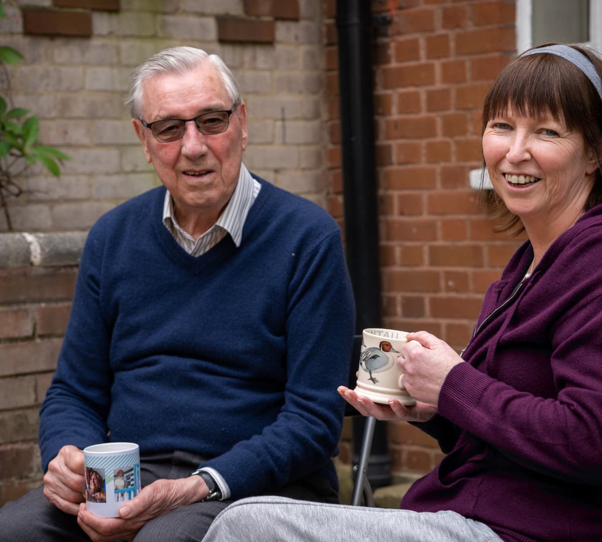 Senior man with grey hair and wearing blue sweatshirt while holding a cup with his carer beside him holding a cup too and both smiling