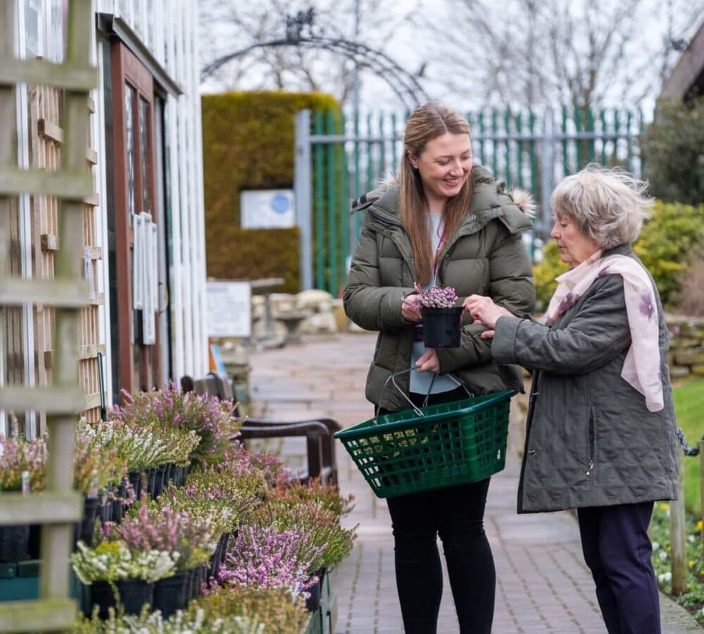 Young female carer with long hair wearing winter clothes and carrying a basket while giving a pot of flowers to an older female adult with grey hair and wearing winter clothes in a flower store