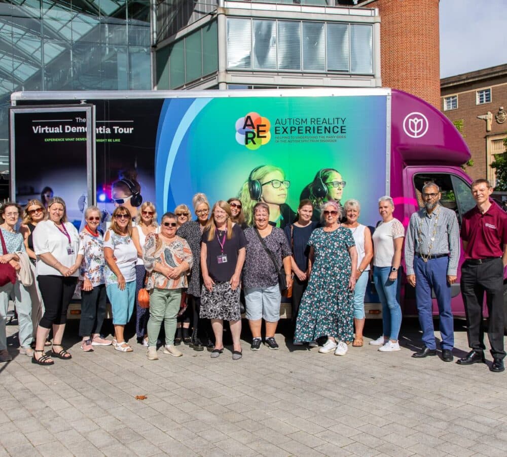 Group of people all happy and smiling and standing in front of the building with a dementia bus at their back
