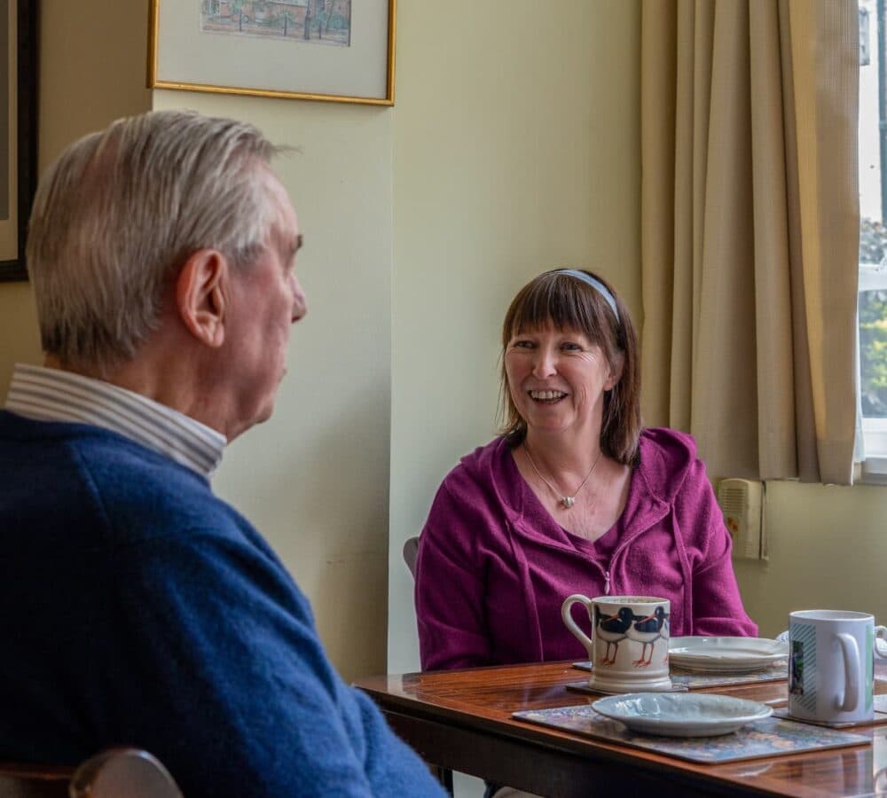 An older man with grey hair sitting on a chair with his young female carer wearing purple happy and smiling while having a cup of tea