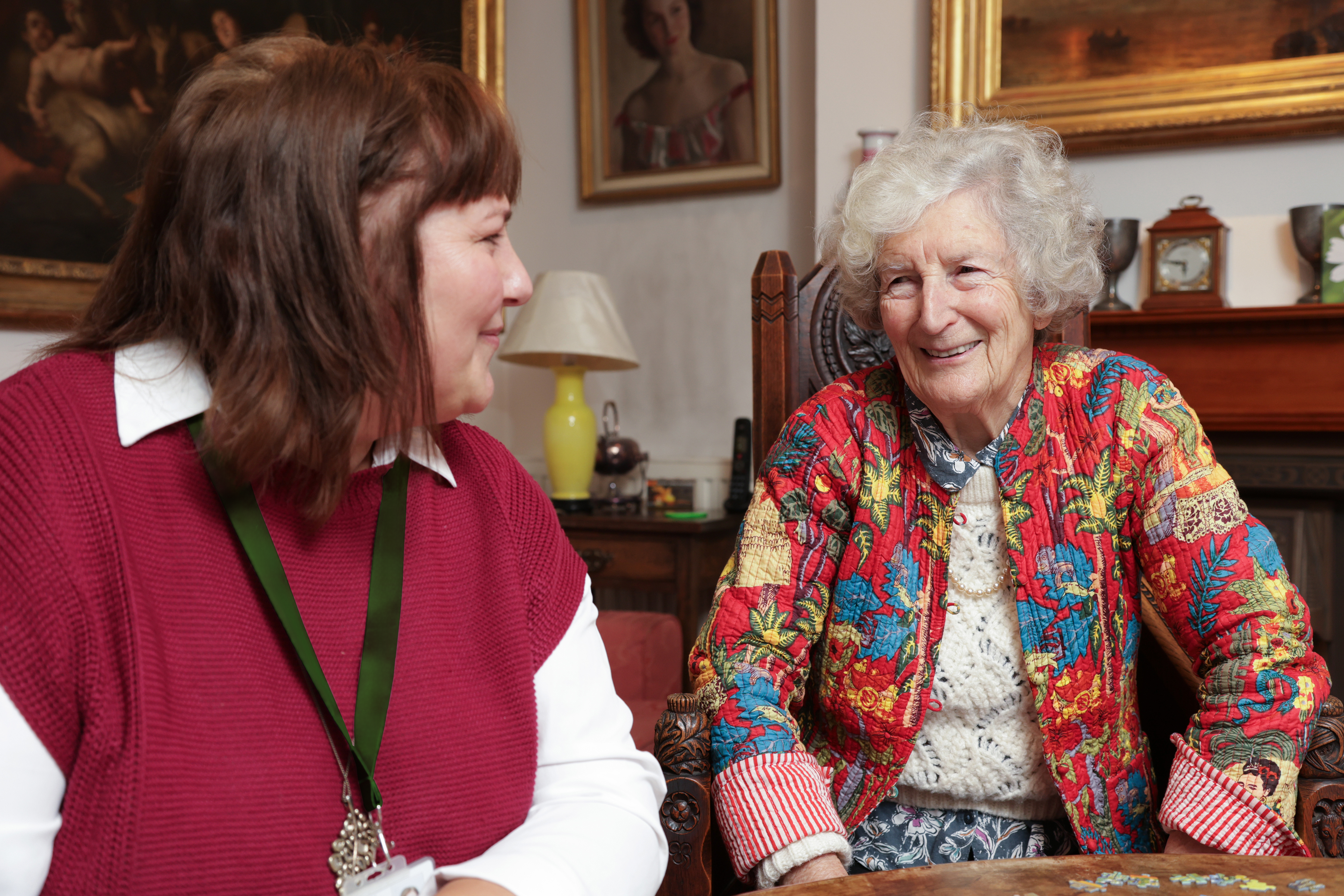 An elderly woman and a caregiver smile and talk while sitting together in a cozy, decorated room. - Home Instead
