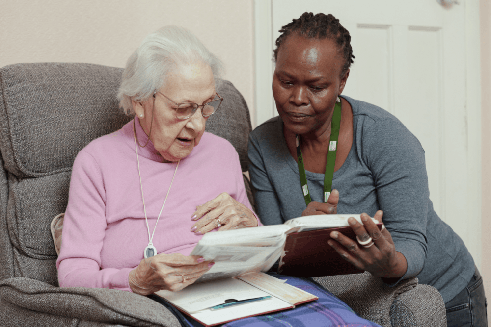 An elderly woman and a caregiver sit together, looking at a photo album in a cozy living room. - Home Instead