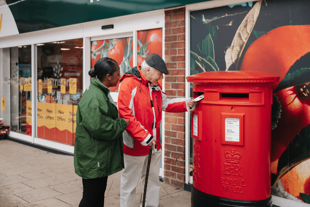 An older gentleman in a flat cap and red jacket is posting a letter into a large Royal Mail postbox outside a supermarket while his female caregiver in a green jacket looks on.