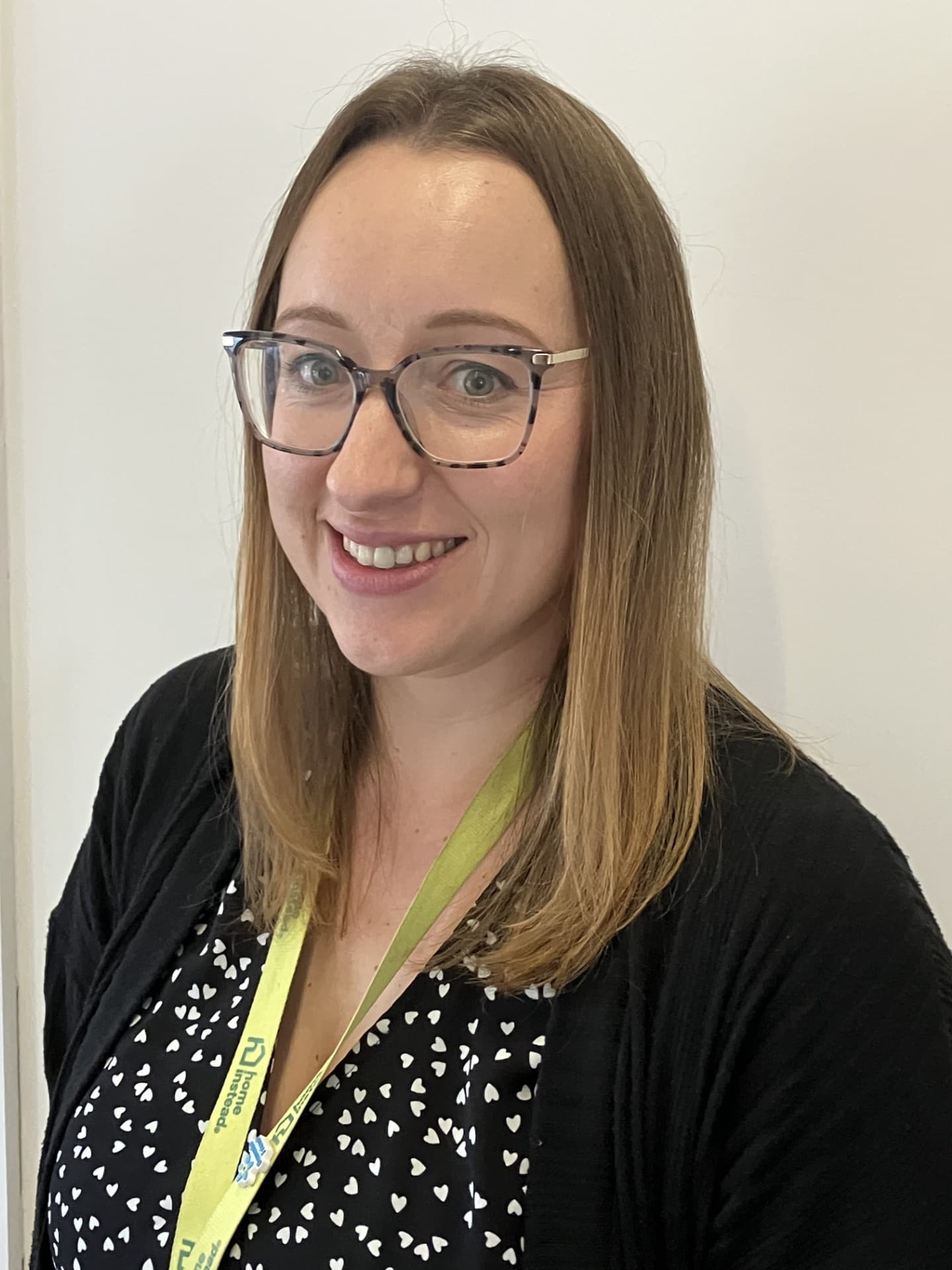 A woman with glasses, wearing a green and black patterned dress, stands against a plain white background. - Home Instead