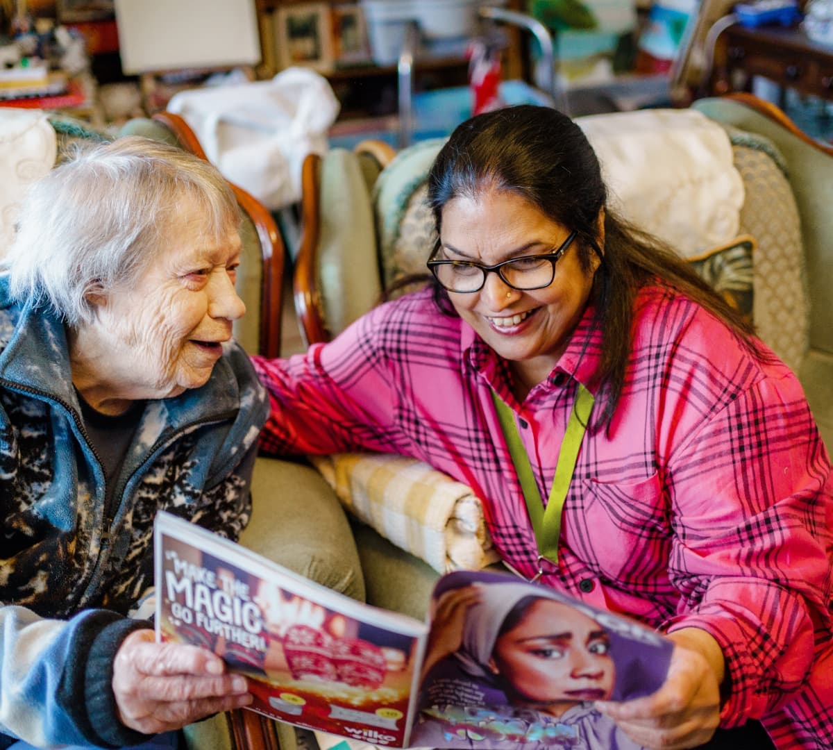 An older woman with grey hair reading together with her younger female carer with long black hair wearing eyeglasses both sitting on a couch inside the house and happily reading together