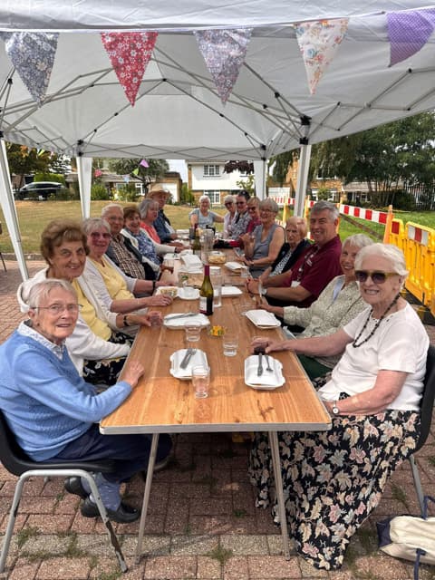 A group of older adults sit around a long table outdoors under a canopy, smiling and enjoying a meal together. - Home Instead