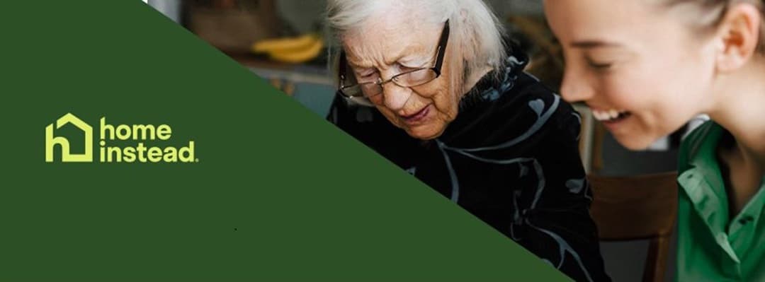 Smiling elderly woman and young carer sitting together, with "Home Instead" logo on a green background. - Home Instead