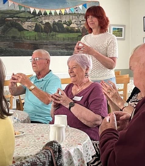 A group of older adults sits and smiles, making hand gestures together in a cheerful indoor setting. - Home Instead