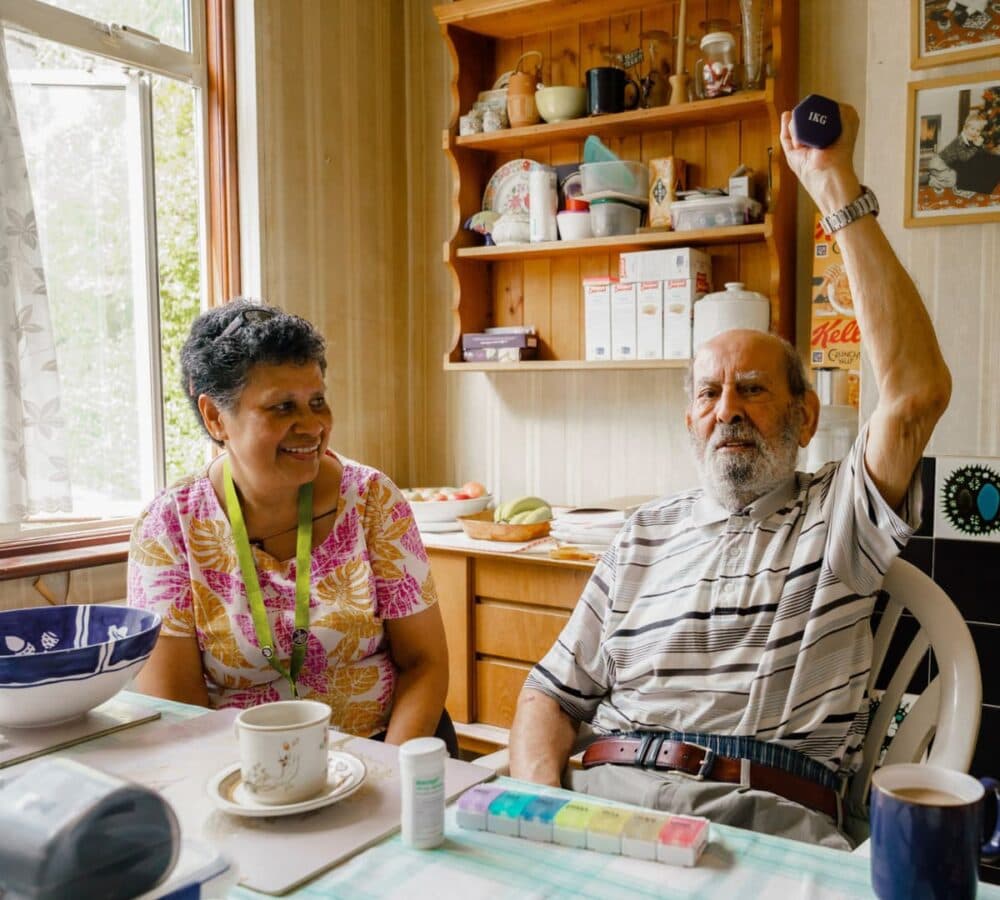 An older male adult exercising with medicines and coffee on the table and while being helped by his younger female carer