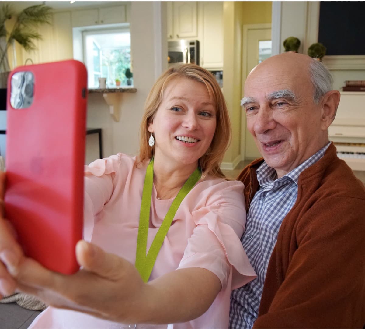 An older male adult with grey hair and wearing sweater and with his younger female carer with long blonde hair and wearing pink both sitting on a couch while taking a selfie
