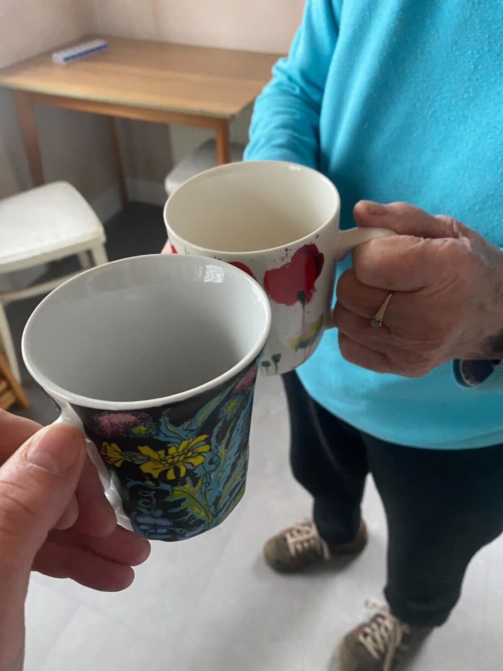Two people clinking colorful mugs together, standing indoors near a wooden table and white chair. - Home Instead