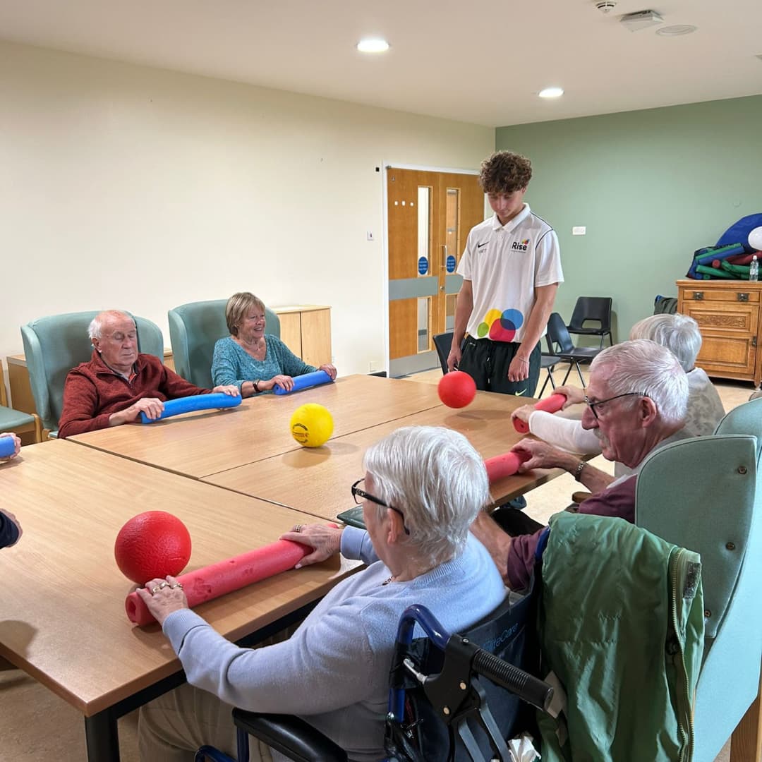 Six seniors play with balls and foam sticks at a table, guided by a young instructor in a care home setting. - Home Instead