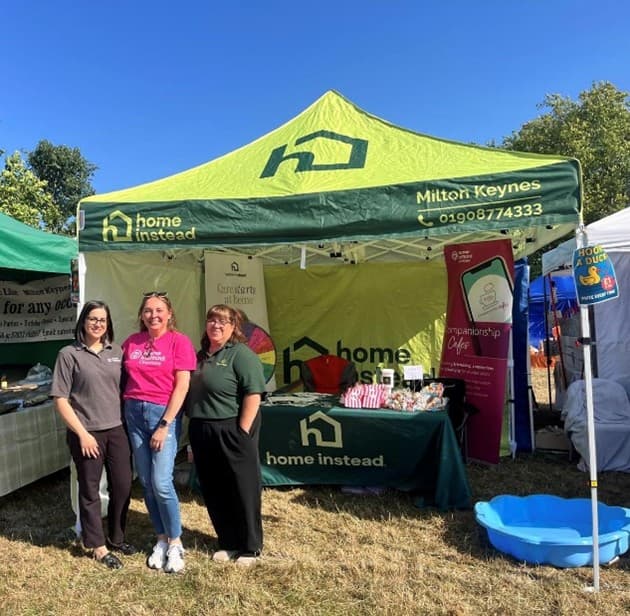 Three women stand smiling in front of a green "Home Instead" tent at an outdoor event on a sunny day. - Home Instead