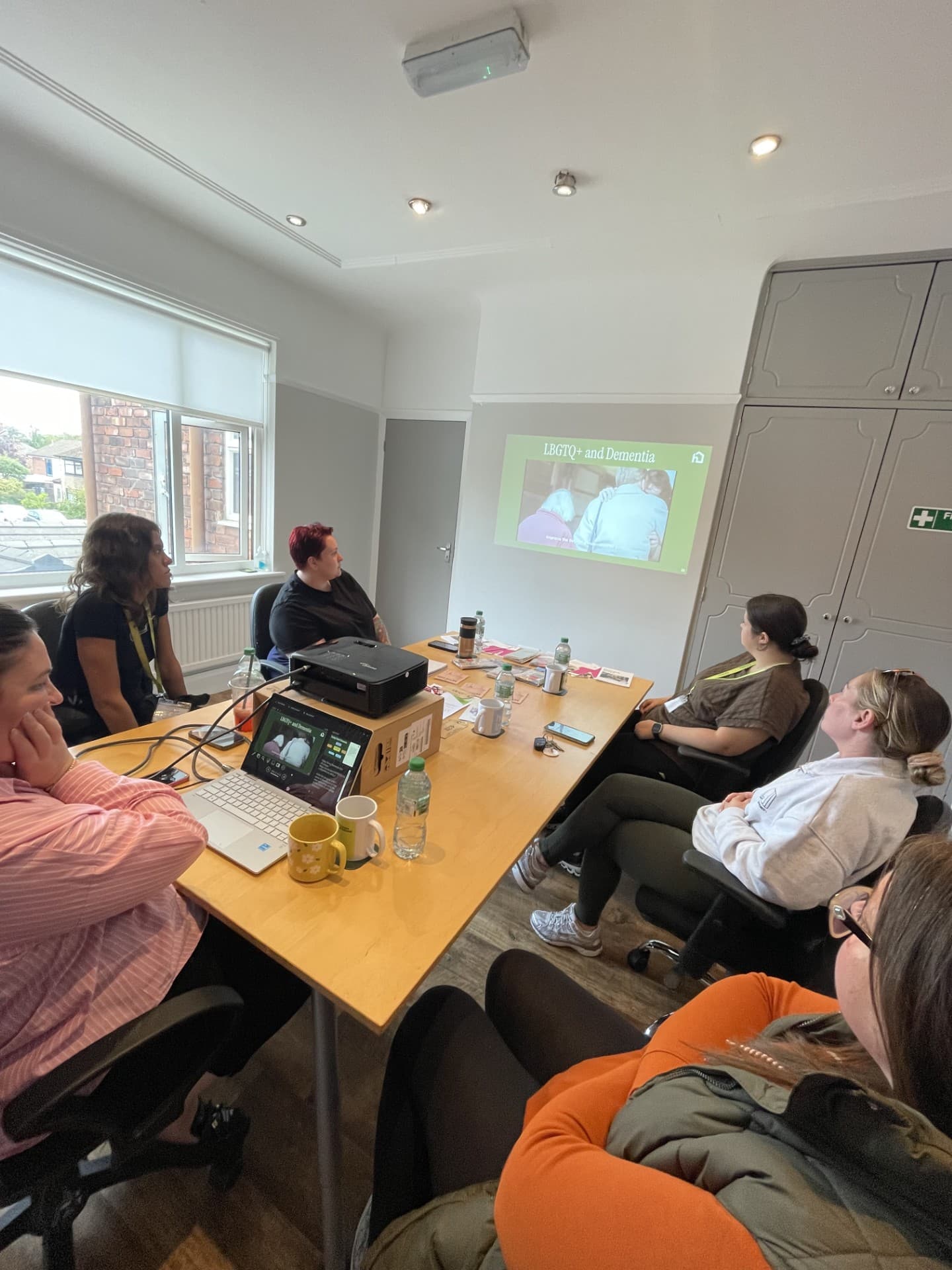 Six people sit around a table watching a presentation on LGBTQ+ and dementia projected on the wall. - Home Instead