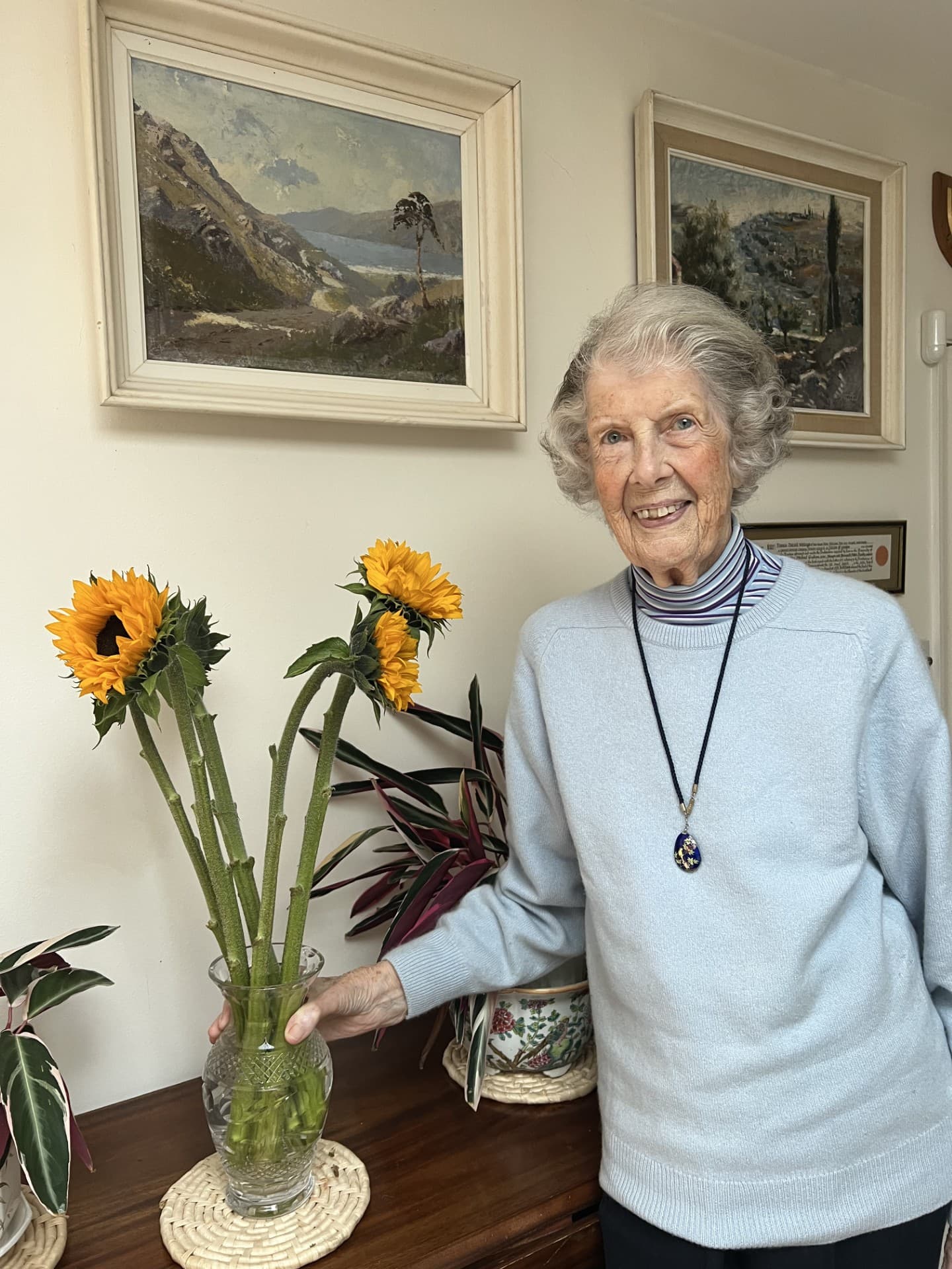 Smiling elderly woman stands beside a vase of sunflowers, with paintings and plants in the background. - Home Instead