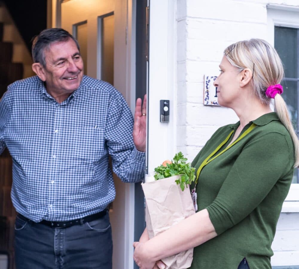 An older male adult at the door welcoming his female carer wearing green and bringing a bag of groceries