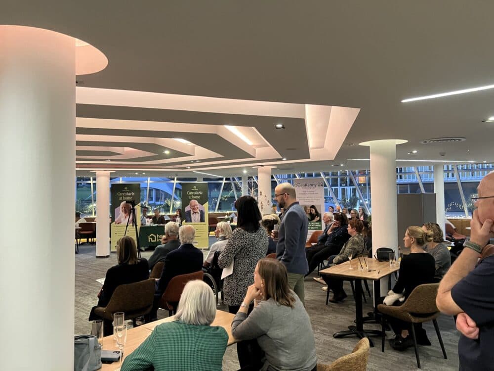 People sit and listen to a speaker at an indoor event with banners and large windows in the background. - Home Instead
