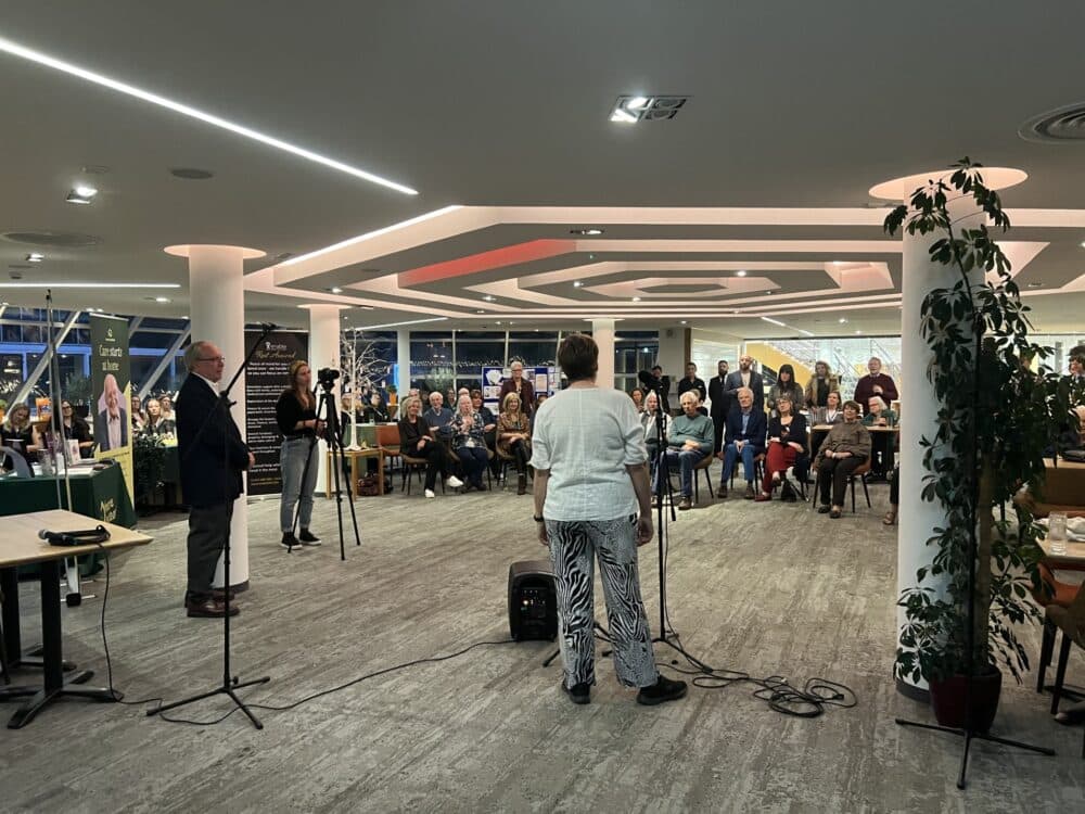 A woman speaks into a microphone facing an audience seated in a modern, well-lit indoor space. - Home Instead