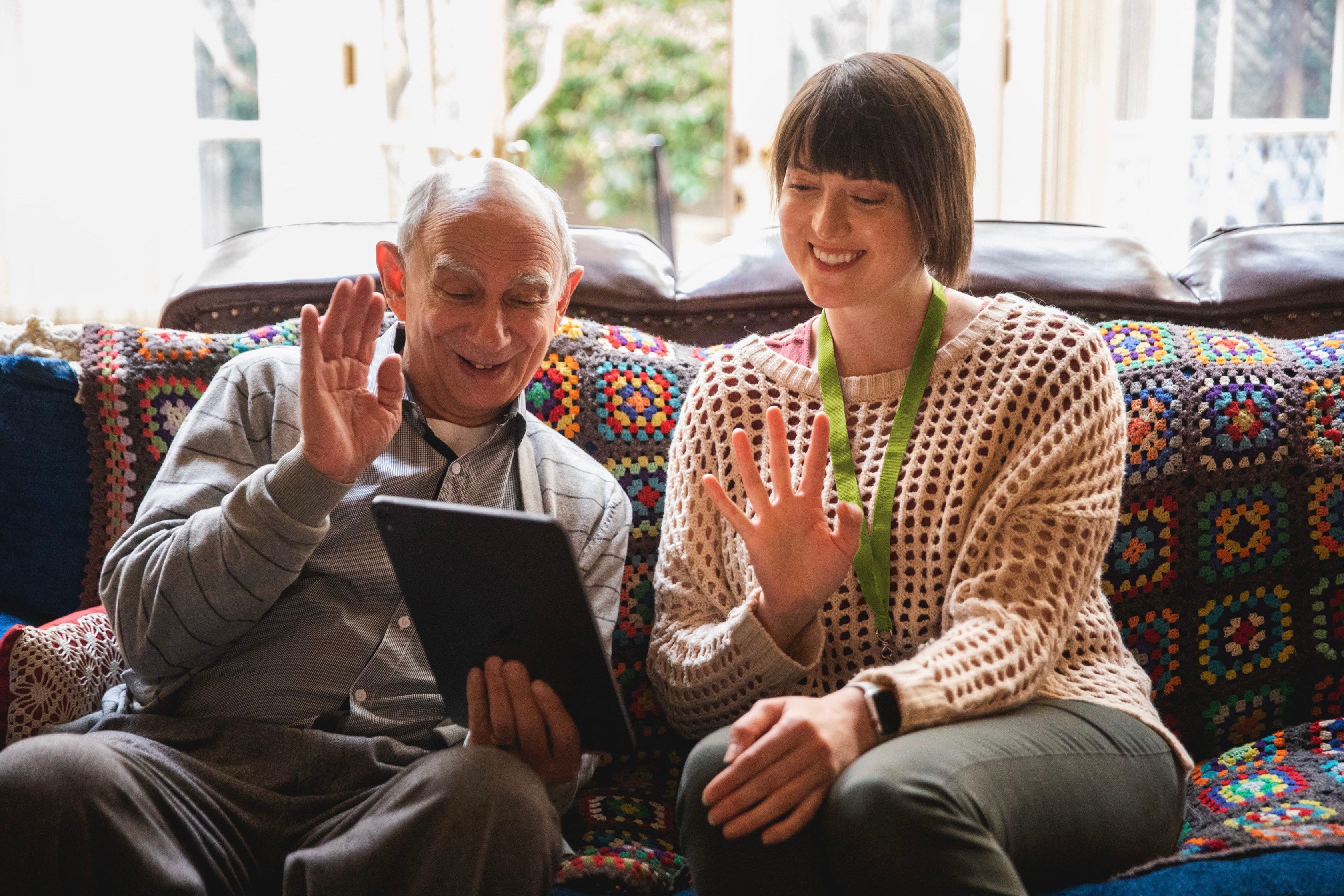 An elderly man and a woman sit on a couch, smiling and waving at a tablet during a video call. - Home Instead