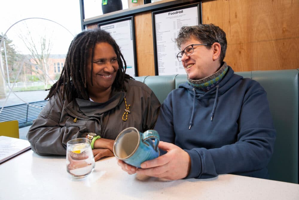 Two people smiling and sitting at a café table, one holding a ceramic mug, with menus in the background. - Home Instead