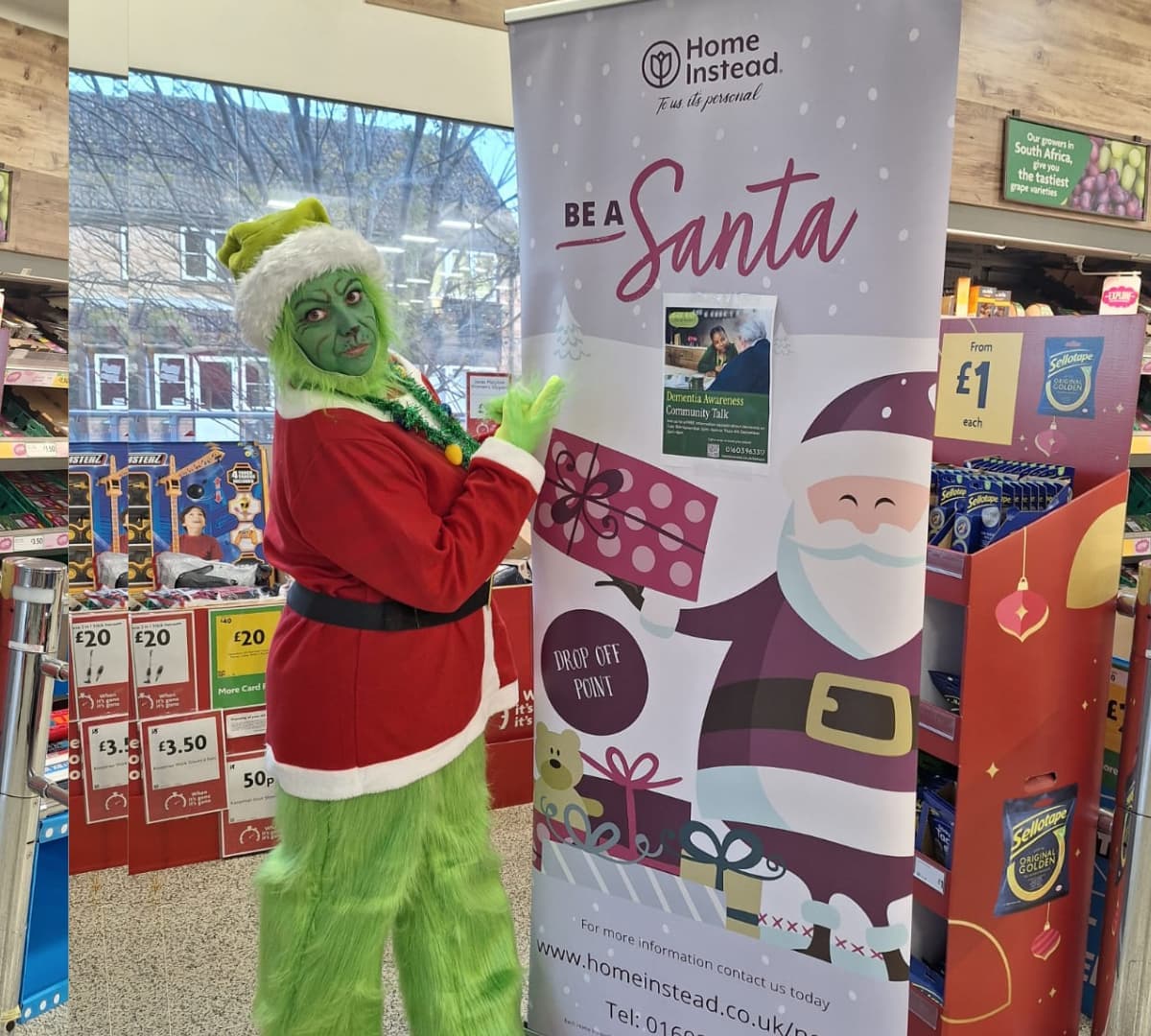 A grinch santa standing beside the banner of a be a santa to a senior