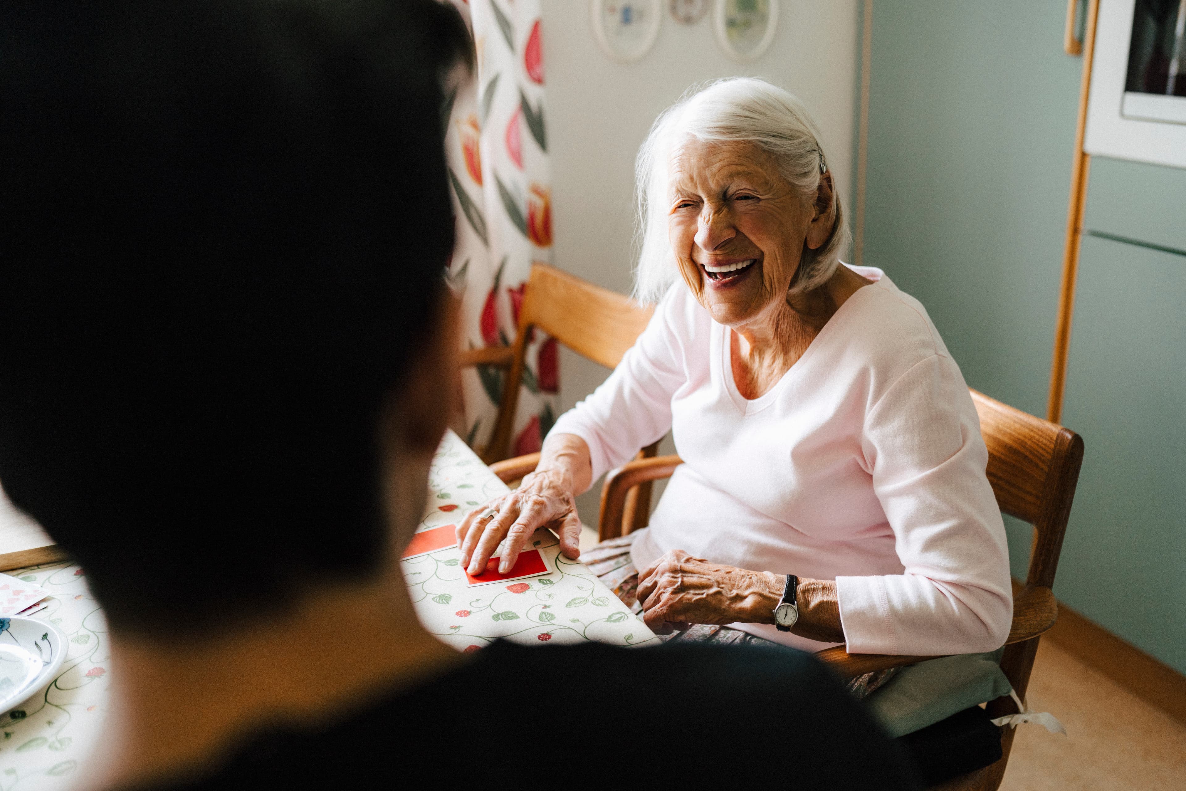 An elderly woman smiles and laughs whilst sitting at a table, talking with someone. - Home Instead