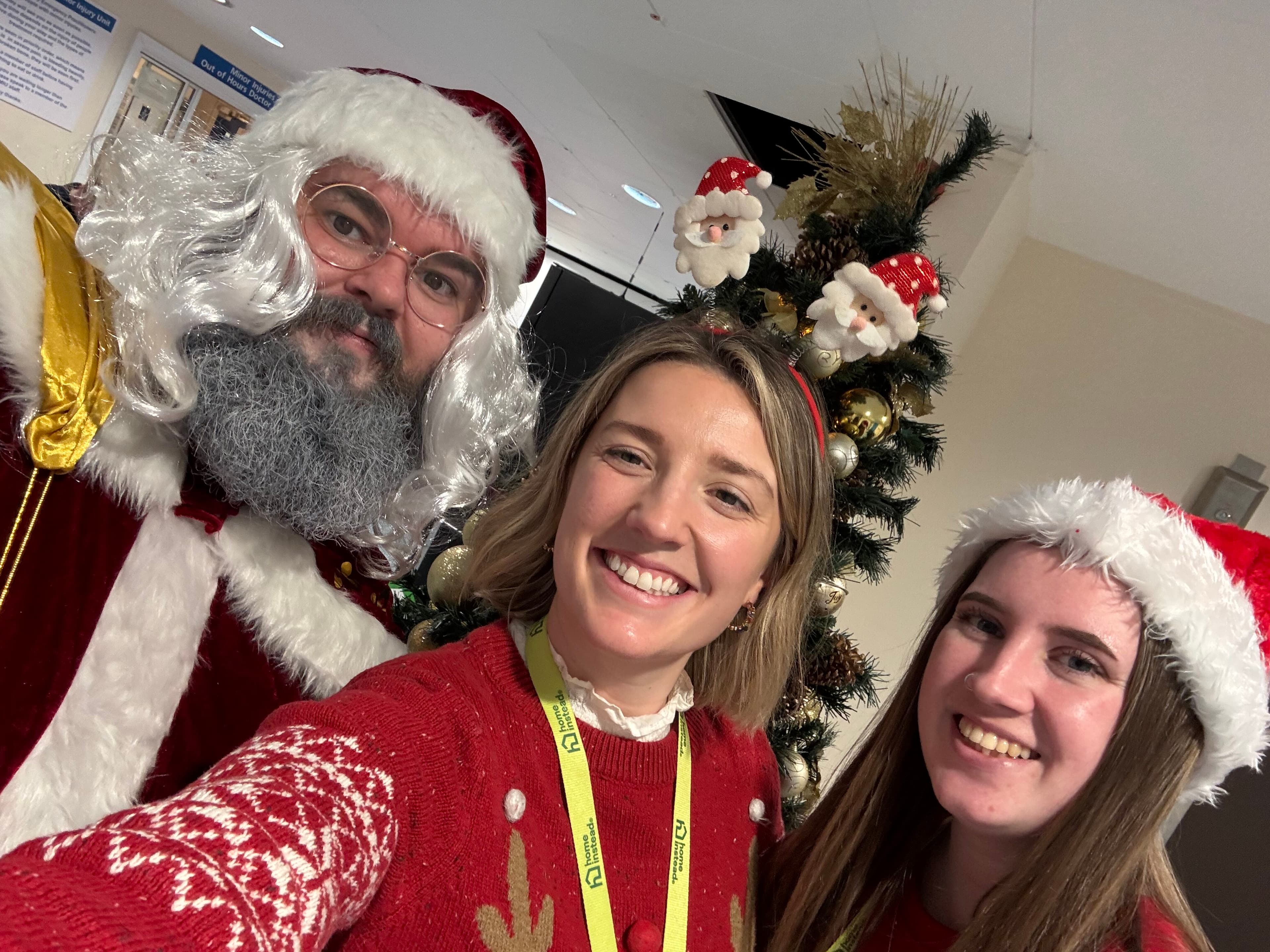Three people in festive attire stand smiling by a decorated Christmas tree with Father Christmas ornaments. - Home Instead