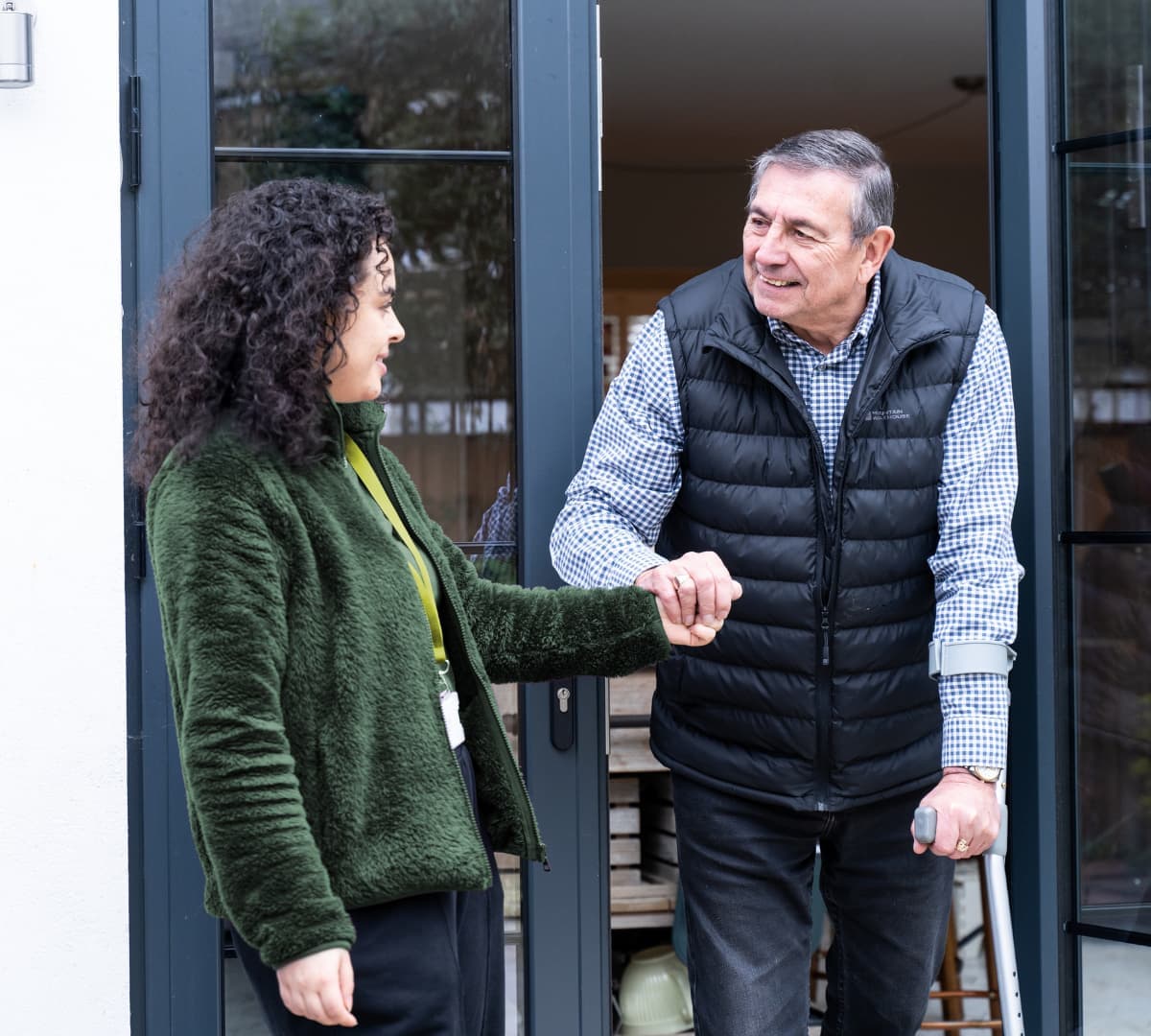 An older male adult wearing a puffer vest and using a crutch while going out of the house and being helped by his younger female carer with curly hair and wearing green