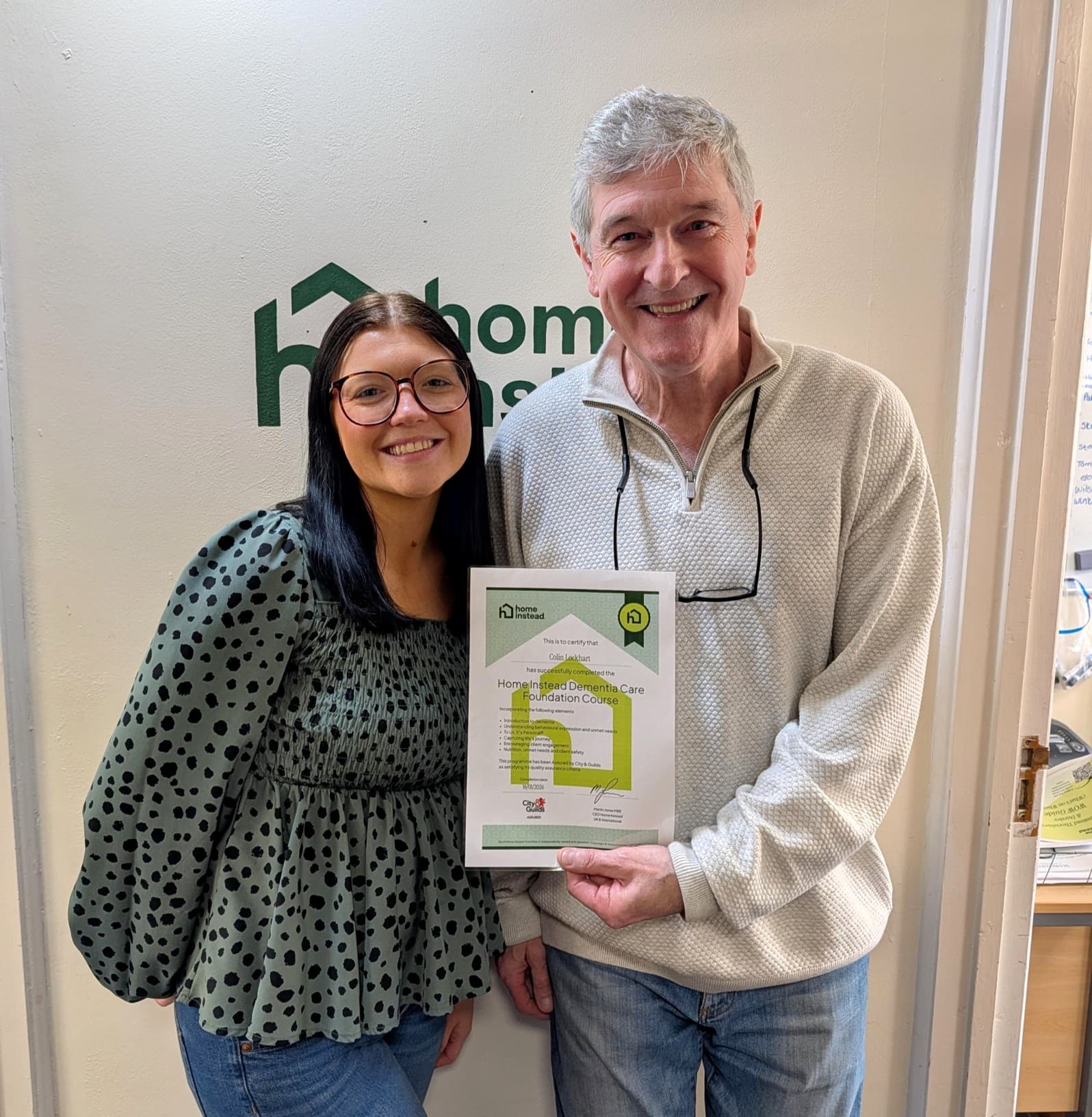 Two people smiling and holding a certificate whilst standing in front of a "home" logo on the wall. - Home Instead
