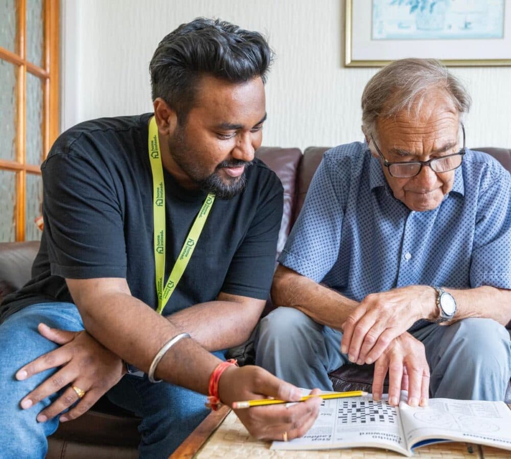 An older male adult wearing eyeglasses and solving a puzzle with his younger male carer with black hair and both sitting on a couch inside the house
