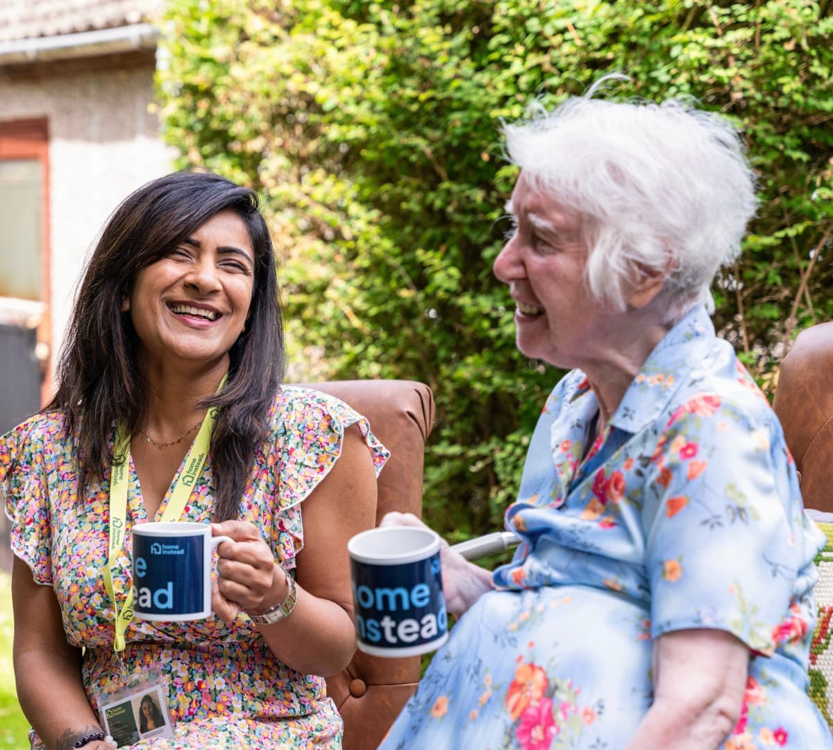 An older female adult with grey hair happy and smiling while sitting and drinking a cup of coffee with her younger female carer in the garden