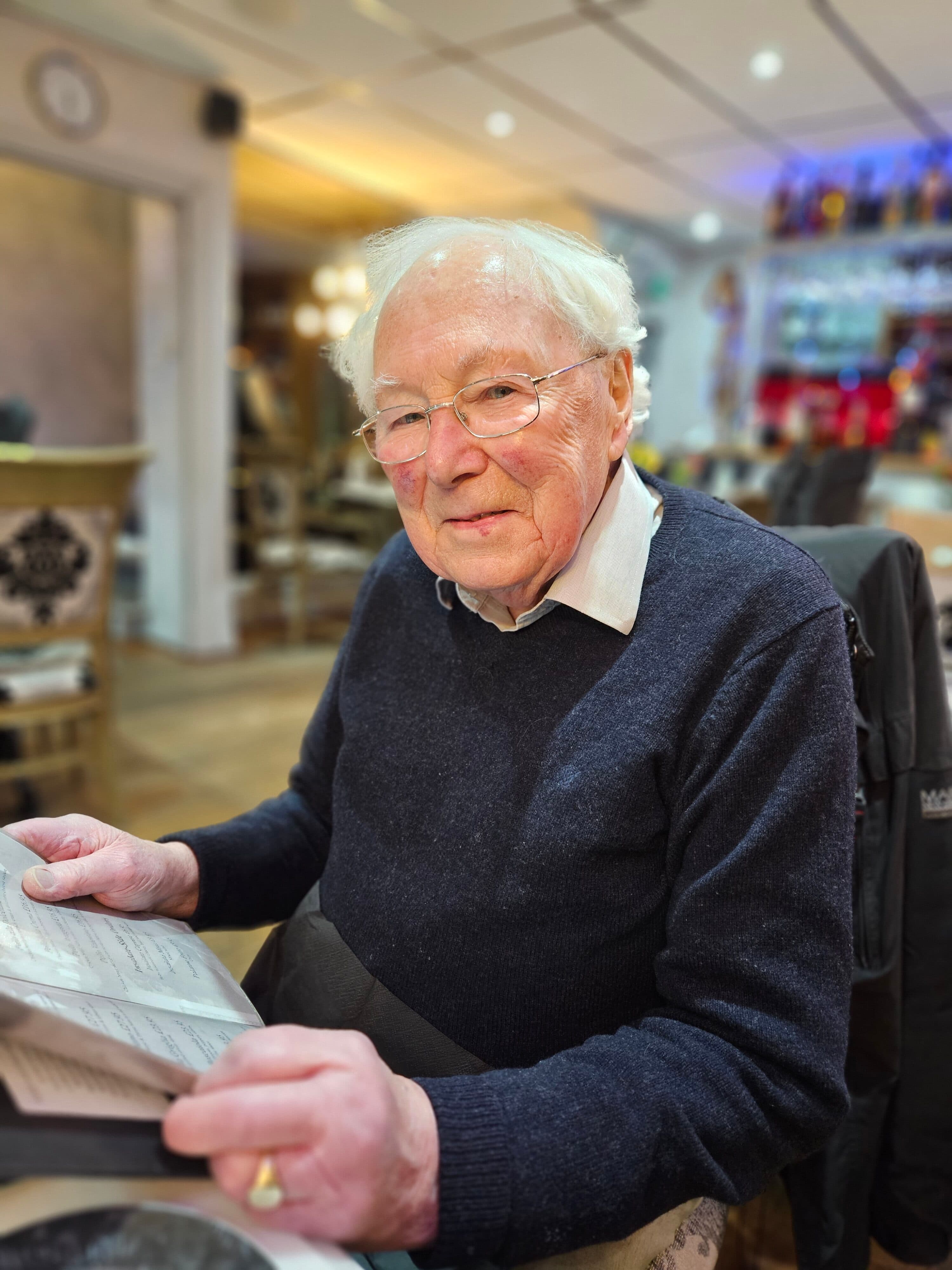 Elderly man with spectacles reading a menu in a warmly lit restaurant, looking at the camera. - Home Instead