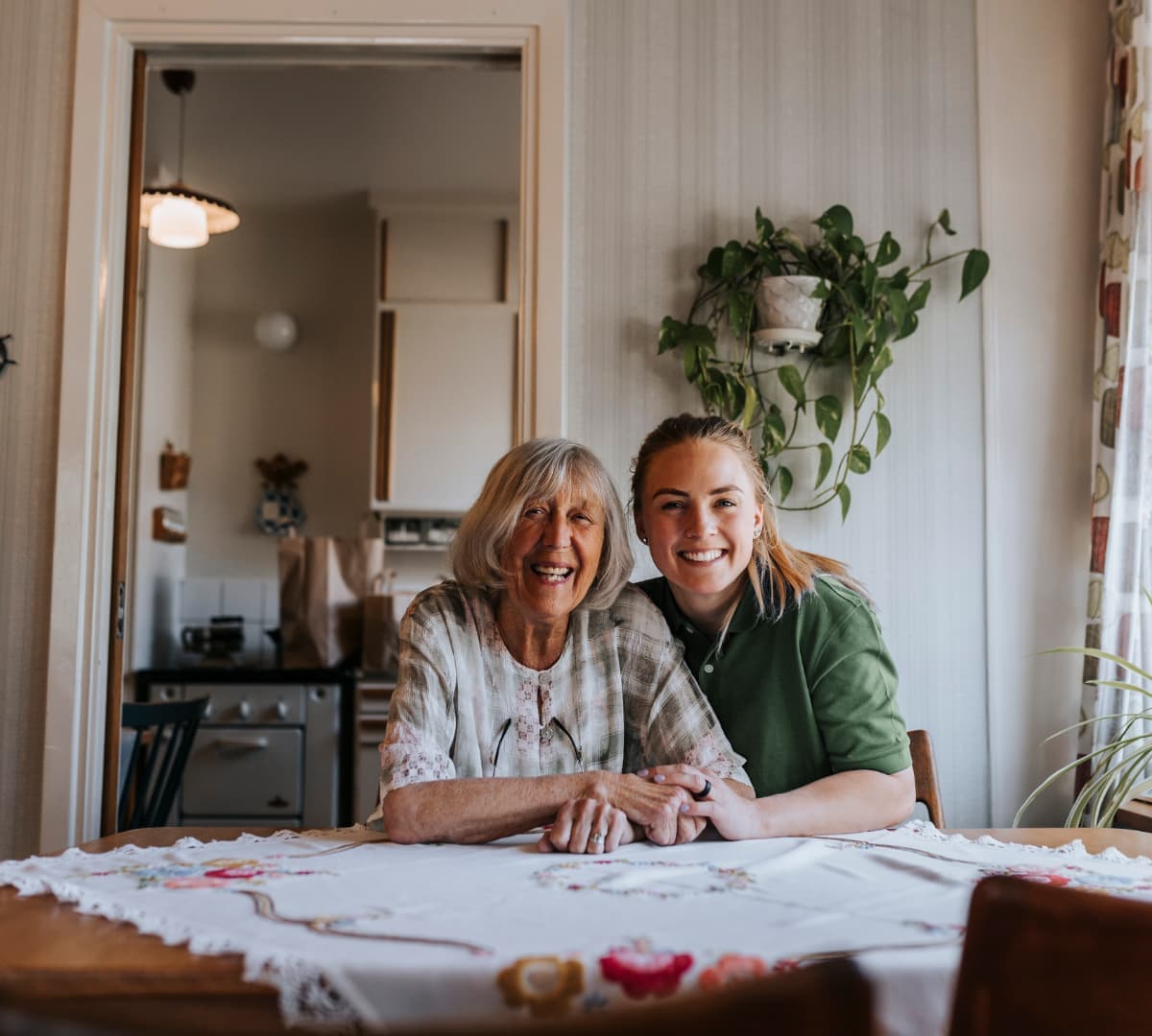 An older female adult with short grey hair happy and smiling while sitting on a table with her younger female carer inside the kitchen