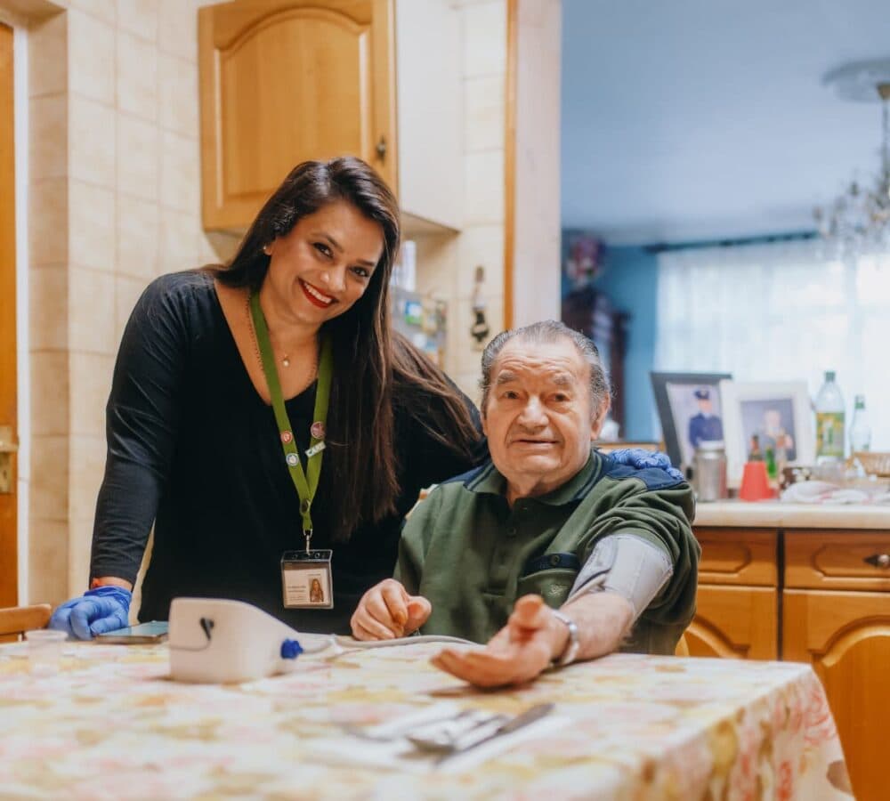 A carer smiles beside an elderly man checking his blood pressure at a kitchen table. - Home Instead