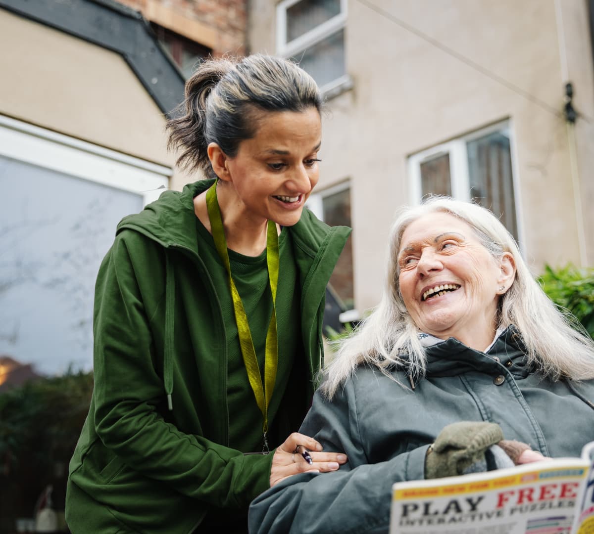 A woman smiles at an older woman in a wheelchair outdoors; both are happy and engaged in conversation. - Home Instead
