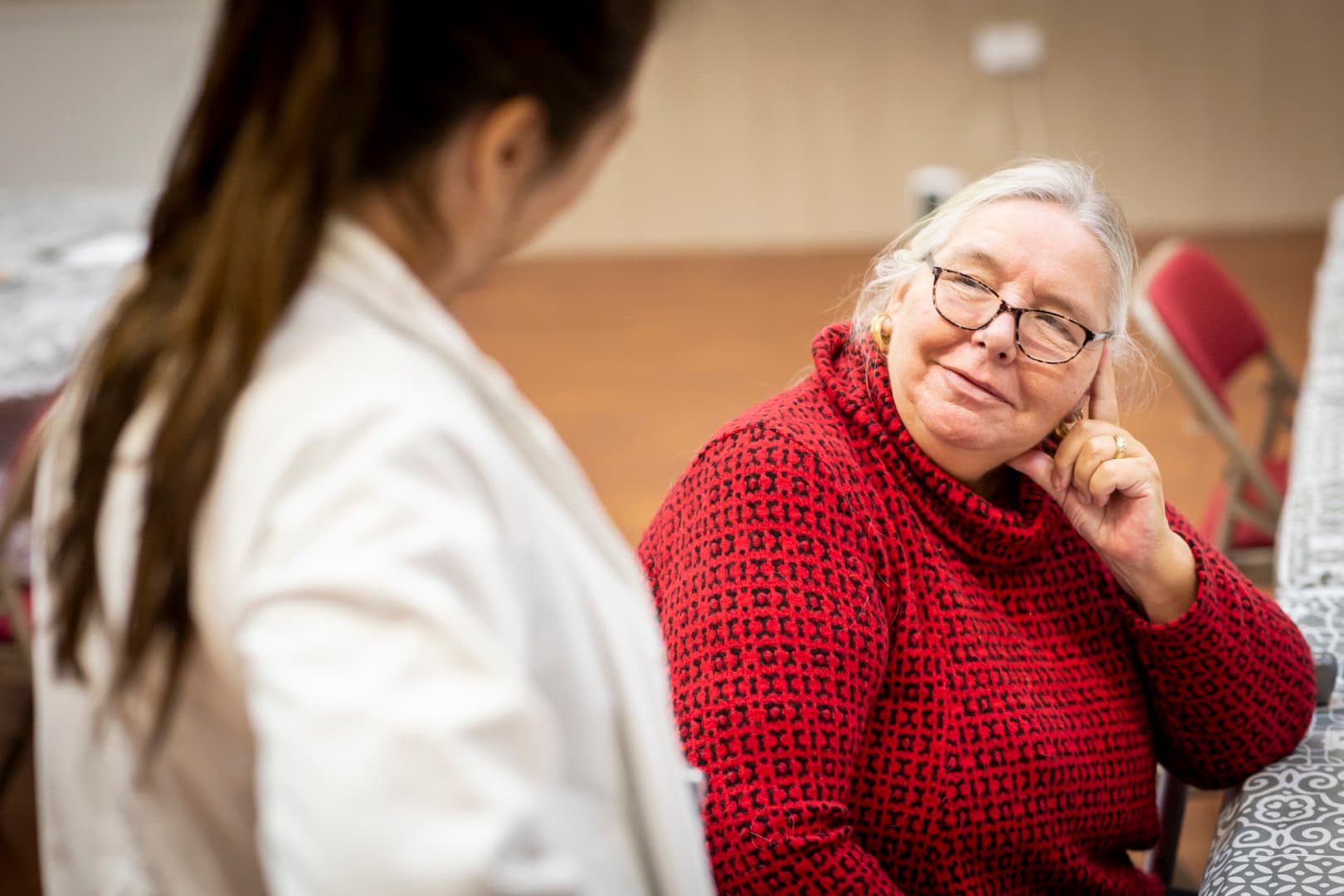 Older woman in red jumper smiles and listens to a person in a white coat during a conversation indoors. - Home Instead