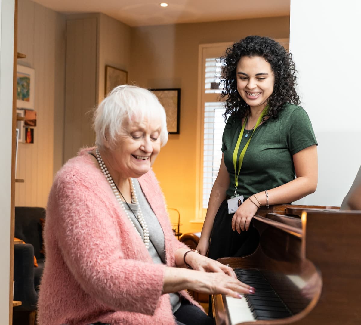 An older female adult with short white hair and wearing pink while playing the piano inside the house with her younger female carer with black curly hair and wearing green both happy and smiling