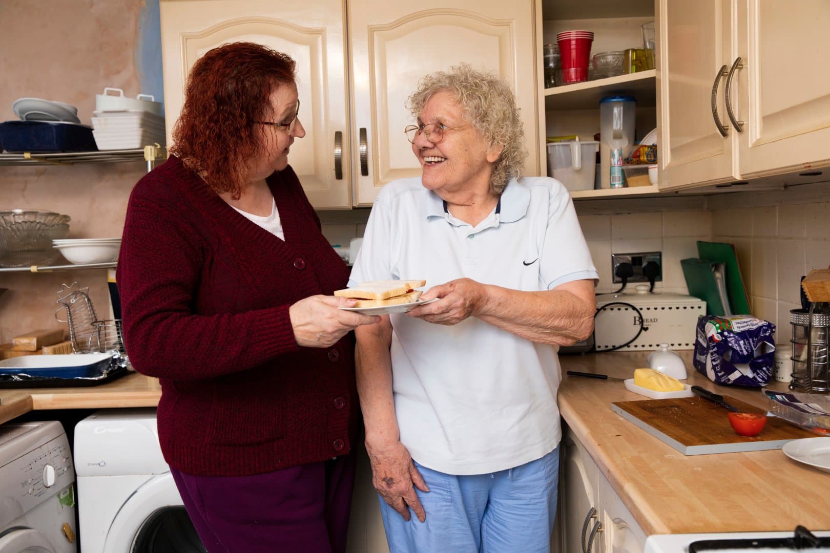 Two smiling women in a kitchen share a plate of sandwiches, standing by the worktop and chatting happily. - Home Instead
