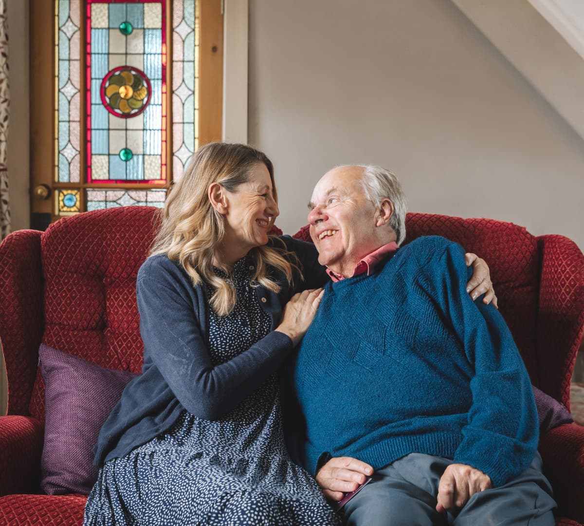 A smiling woman and older man sit together on a red sofa, sharing a warm, affectionate moment at home. - Home Instead