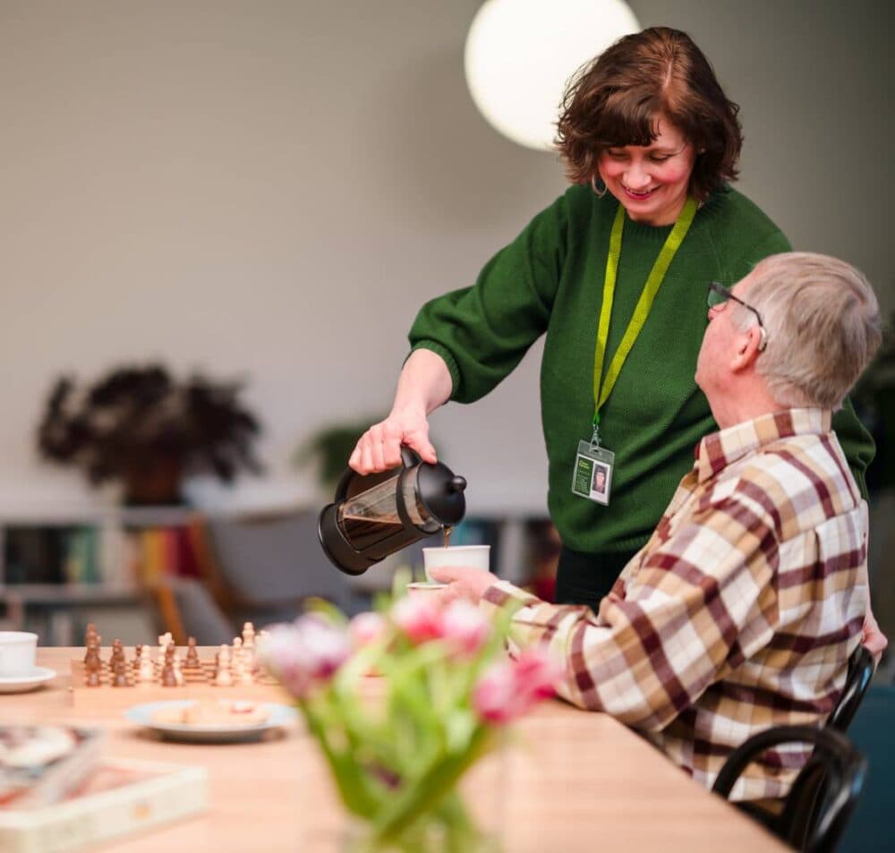 A woman pours coffee for an elderly man at a table with chess pieces and flowers. - Home Instead
