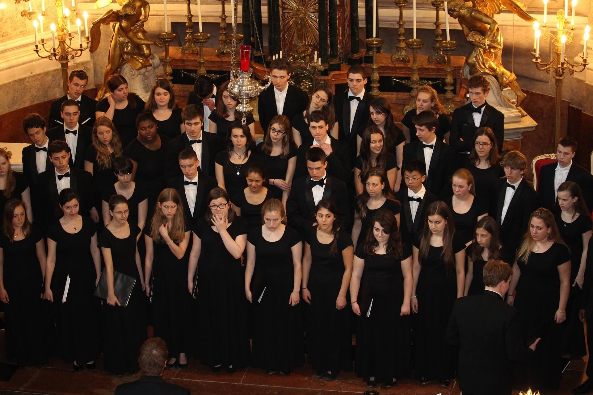 A choir dressed in black attire performs in an ornate setting with chandeliers and candles, facing a conductor. - Home Instead