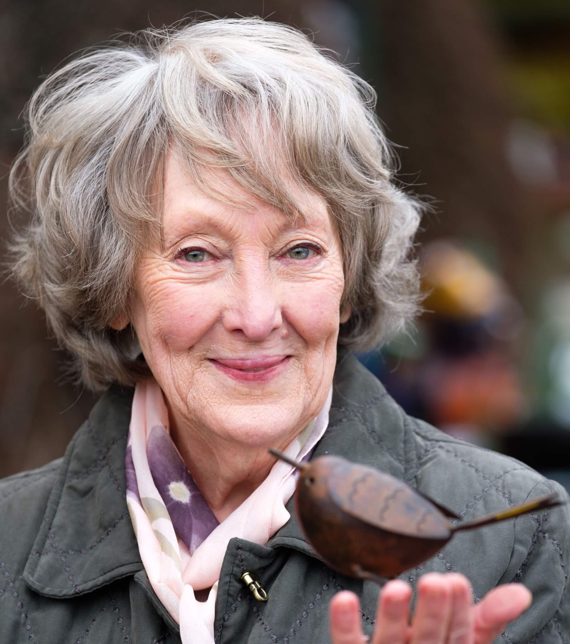 A smiling elderly woman with gray hair holds a small bird sculpture in her hand, wearing a scarf and a jacket. - Home Instead