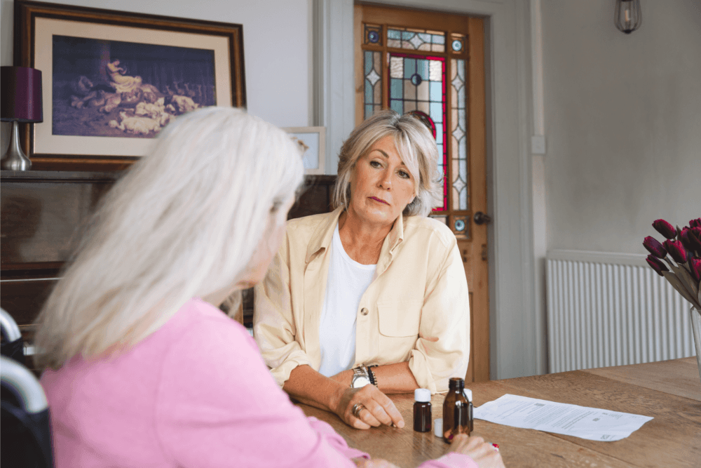 Two older women sitting at a table, discussing something serious; one is holding a paper, with medicine bottles on the table. - Home Instead