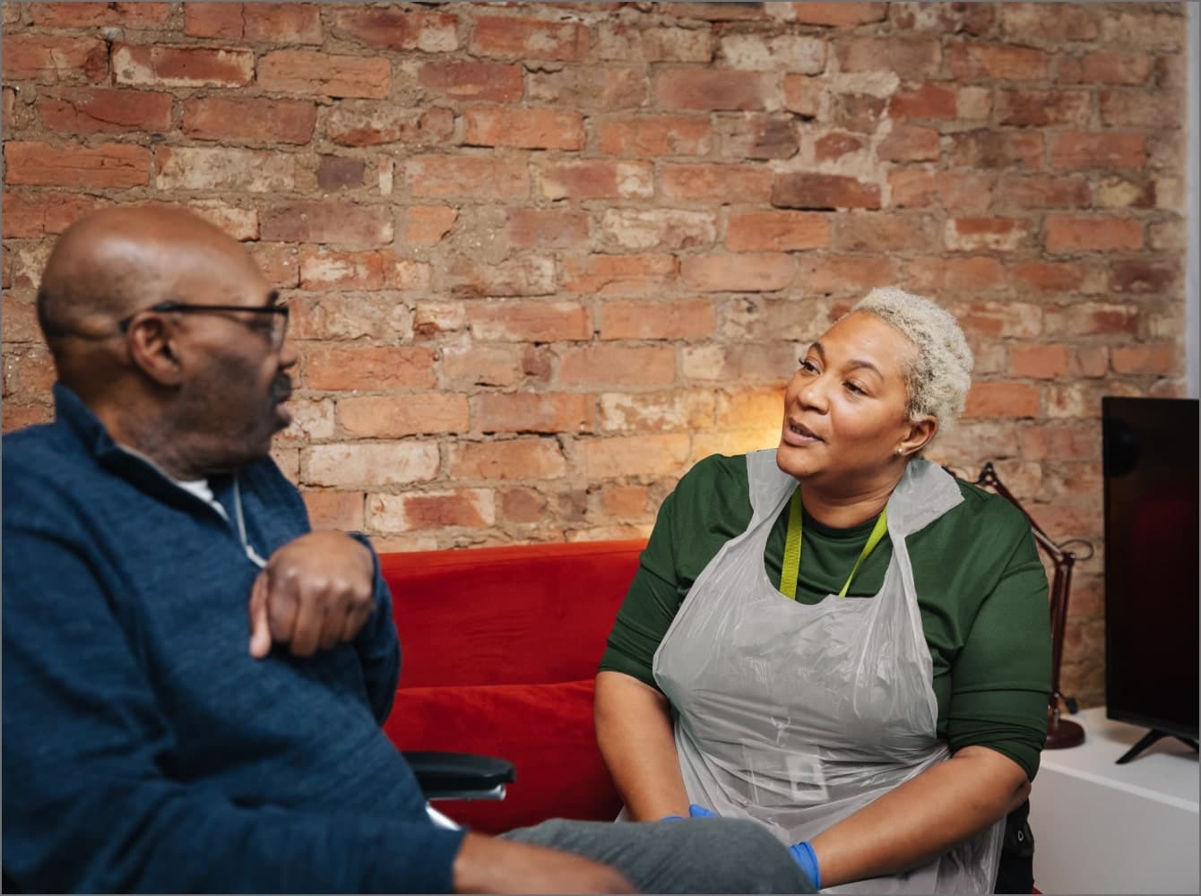 A woman with a name badge smiles while petting a dog, sitting with an older man on a gray couch in a bright room. - Home Instead